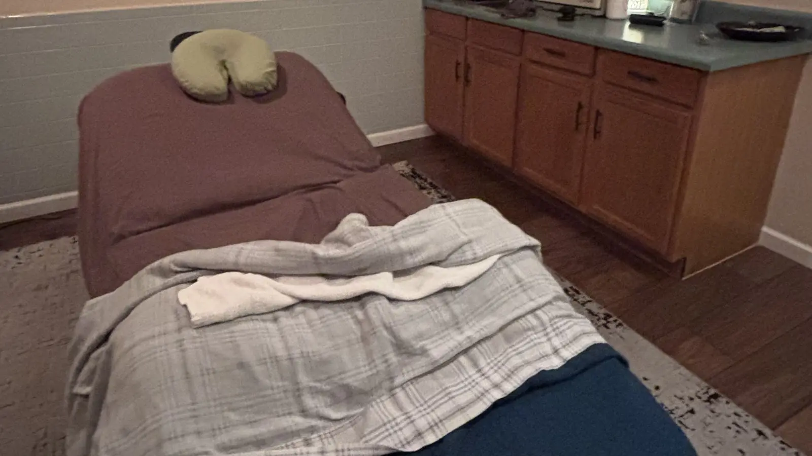 Treatment room with a plum-sheeted massage table, green face cradle, a plaid blanket, teal counter and wood cabinets